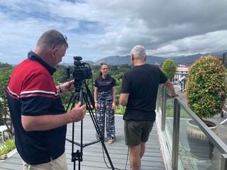 A cameraman filming a woman on a deck against a backdrop of grass and mountains