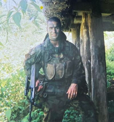 Jared in an Army uniform hold a firearm at his side in a jungle