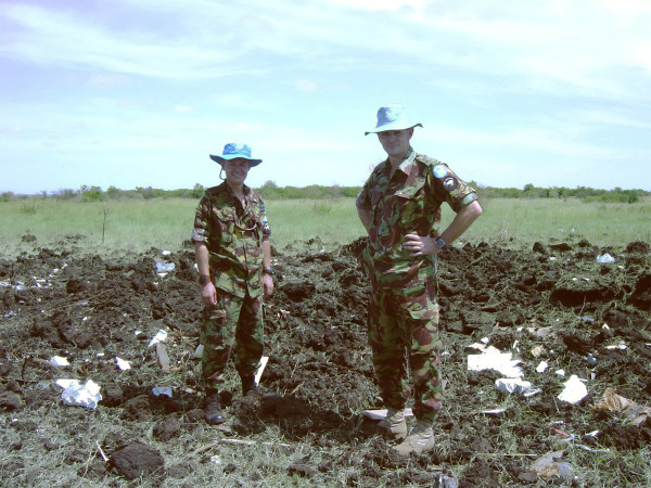 Two men in army uniforms standing in a field of upturned soil and debris. 