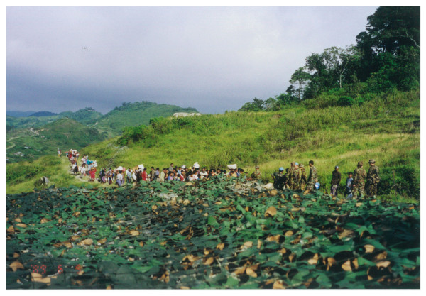 A line of people walking through grassy hills with a military escort.