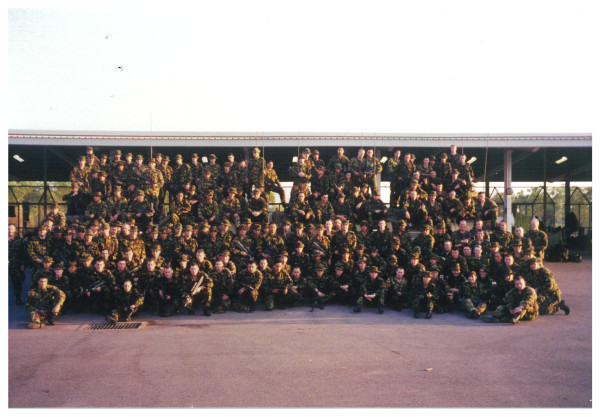 Approximately 100 soldiers in army uniforms posing for a photo on a concrete ground.